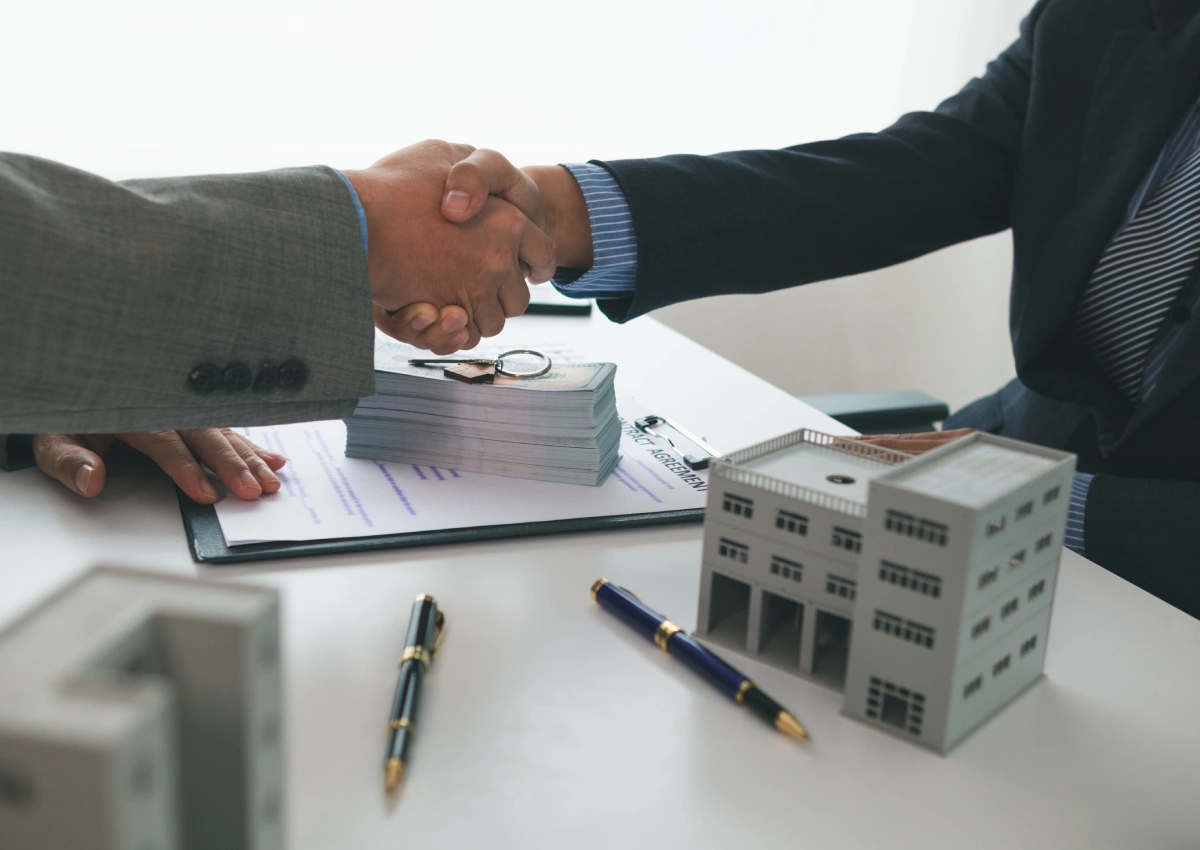 Two people shaking hands over a table with pens and papers.